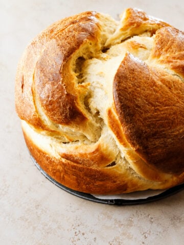 A round loaf of golden-brown, fluffy Cinnamon Raisin Round Challah Bread with a cracked top sits on a light surface, showcasing its soft, airy texture and shiny crust.