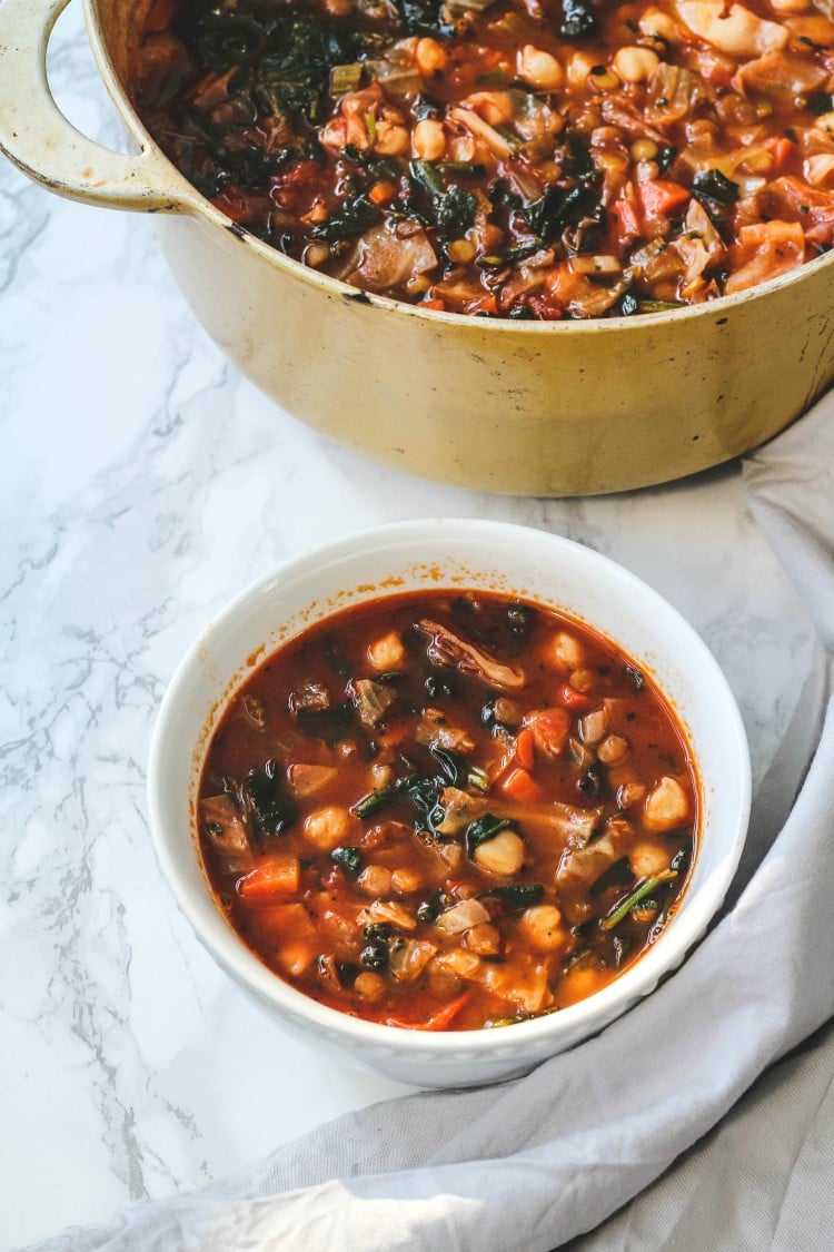 close up image of cabbage and lentil soup in a bowl and dutch oven on a marble background.