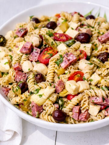 A large white bowl filled with Antipasto Pasta Salad-rotini pasta mixed with cherry tomatoes, mozzarella balls, salami slices, black olives, chopped herbs, and pickled vegetables.