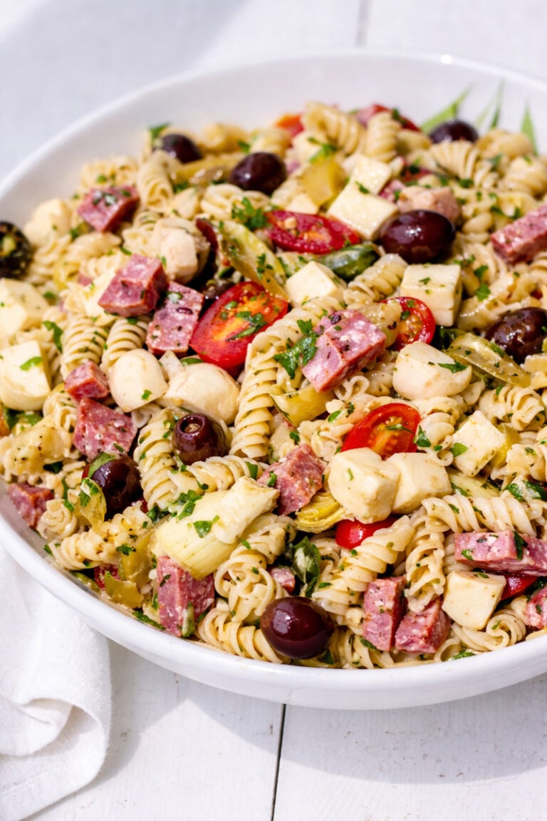 A large white bowl filled with Antipasto Pasta Salad—rotini pasta mixed with cherry tomatoes, mozzarella balls, salami slices, black olives, chopped herbs, and pickled vegetables.