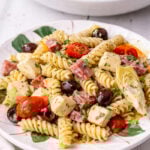 A plate of Antipasto Pasta Salad with rotini, cherry tomatoes, black olives, artichoke hearts, cubes of cheese, and slices of salami, garnished with chopped herbs. Another plate of the same salad is in the background.