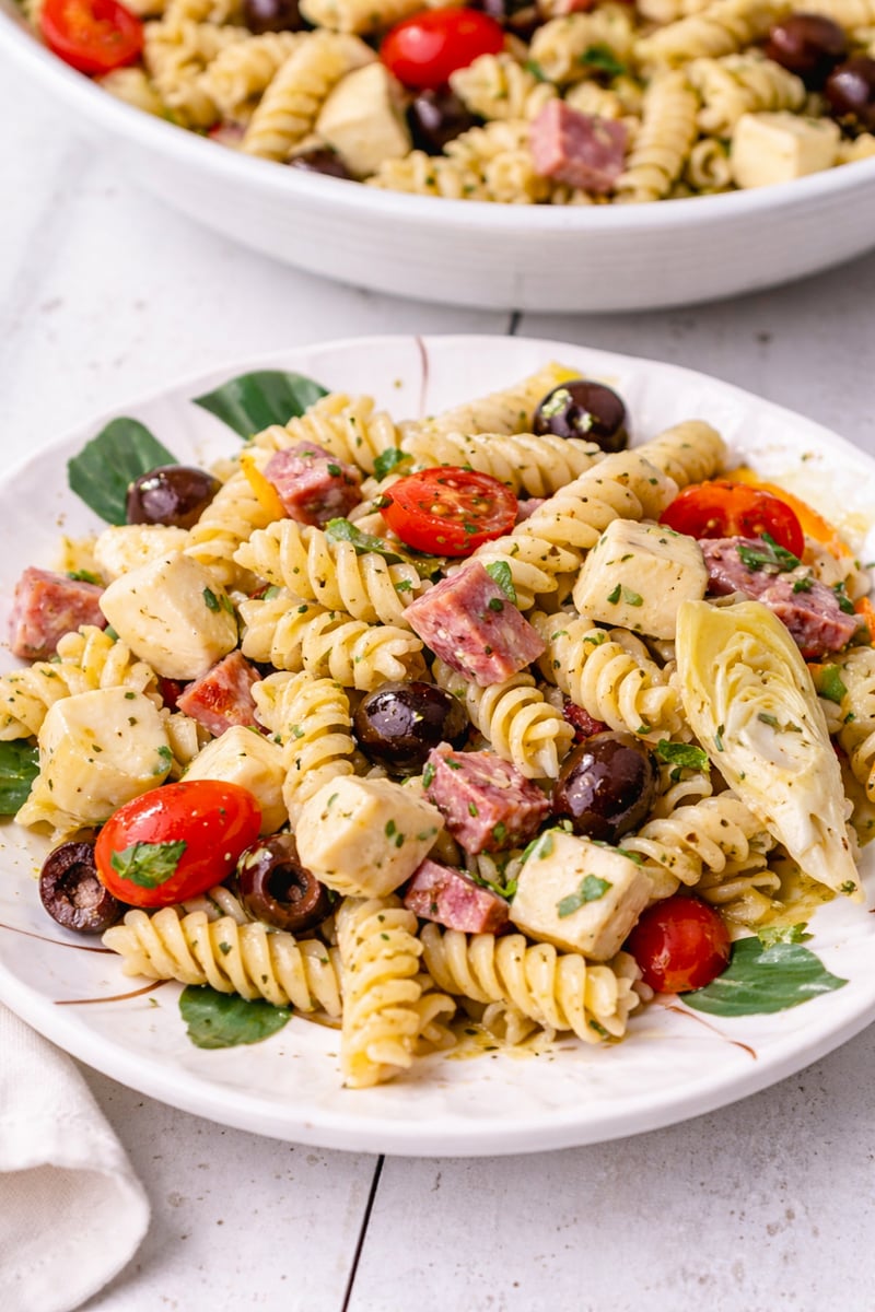 A plate of Antipasto Pasta Salad with rotini, cherry tomatoes, black olives, artichoke hearts, cubes of cheese, and slices of salami, garnished with chopped herbs. Another plate of the same salad is in the background.