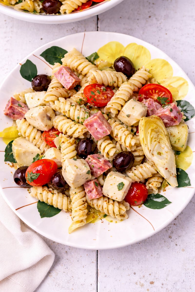 A plate of Antipasto Pasta Salad with rotini, cherry tomatoes, mozzarella cubes, black olives, salami pieces, artichoke hearts, and fresh herbs, arranged on a white plate with yellow flower accents.