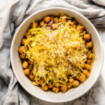 A white bowl of Cacio e Pepe Spaghetti Squash with Chickpeas, featuring cooked chickpeas and shredded spaghetti squash topped with breadcrumbs and seasonings, sits on a marble surface next to a gray cloth.