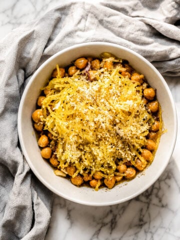 A white bowl of Cacio e Pepe Spaghetti Squash with Chickpeas, featuring cooked chickpeas and shredded spaghetti squash topped with breadcrumbs and seasonings, sits on a marble surface next to a gray cloth.