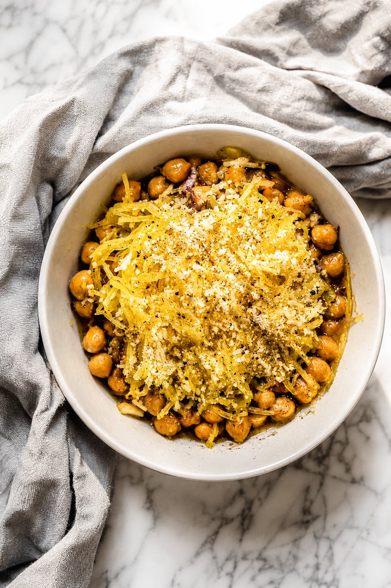 A white bowl of Cacio e Pepe Spaghetti Squash with Chickpeas, featuring cooked chickpeas and shredded spaghetti squash topped with breadcrumbs and seasonings, sits on a marble surface next to a gray cloth.