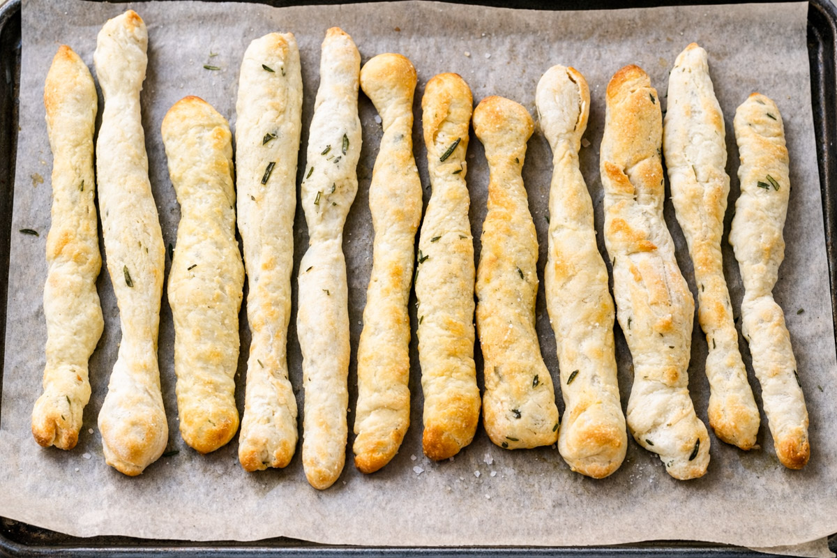 Twelve golden-brown Rosemary Grissini with a slightly twisted shape are lined up on a baking tray covered with parchment paper. Some breadsticks have small bits of herbs baked into them.