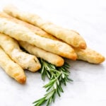 Six golden-brown Rosemary Grissini are stacked on a white marble surface, with a small sprig of fresh rosemary placed underneath.