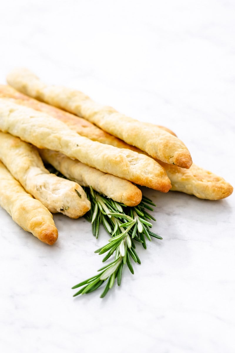 Six golden-brown Rosemary Grissini are stacked on a white marble surface, with a small sprig of fresh rosemary placed underneath.