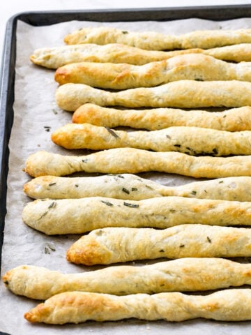 A baking sheet lined with parchment paper holds several golden-brown, twisted Rosemary Grissini sprinkled with herbs.