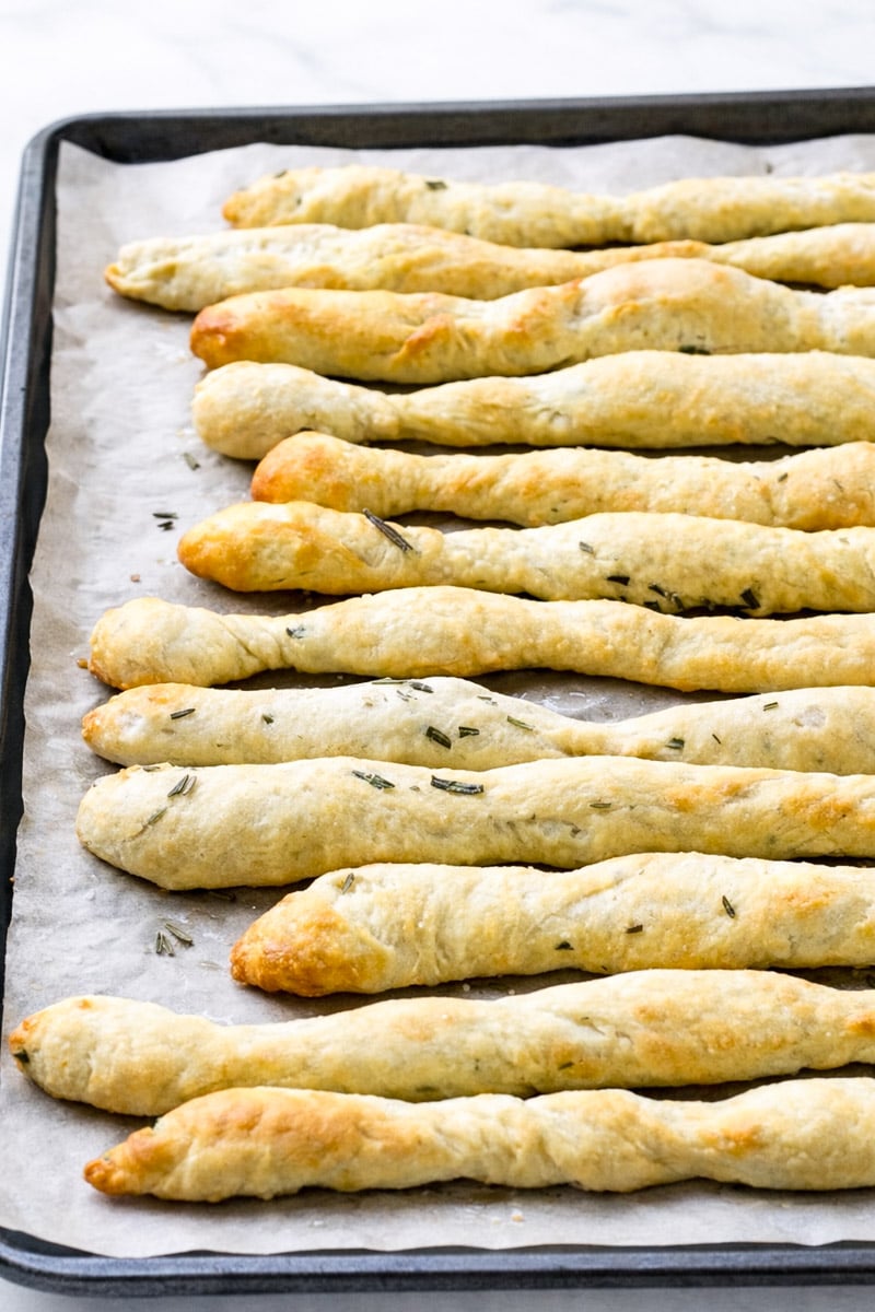 A baking sheet lined with parchment paper holds several golden-brown, twisted Rosemary Grissini sprinkled with herbs.