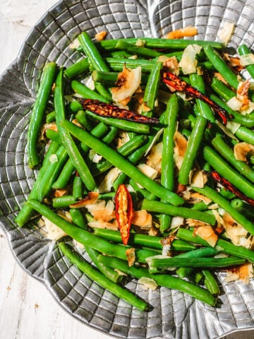Caramelized coconut green beans tossed with toasted coconut flakes and dried red chili peppers in a textured silver bowl.