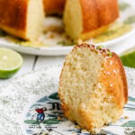 A slice of golden Coconut Lime Bundt Cake with delicate coconut flakes sits on a decorative plate, while the rest of the cake and a halved lime tempt in the background.