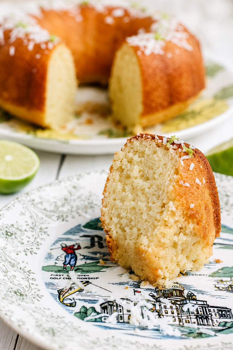 A slice of golden Coconut Lime Bundt Cake with delicate coconut flakes sits on a decorative plate, while the rest of the cake and a halved lime tempt in the background.