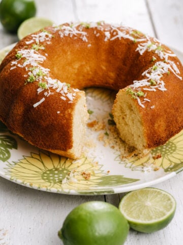 A Coconut Lime Bundt Cake topped with shredded coconut and lime zest sits on a floral plate. A slice is missing, and fresh limes are placed beside the cake on a white wooden surface.