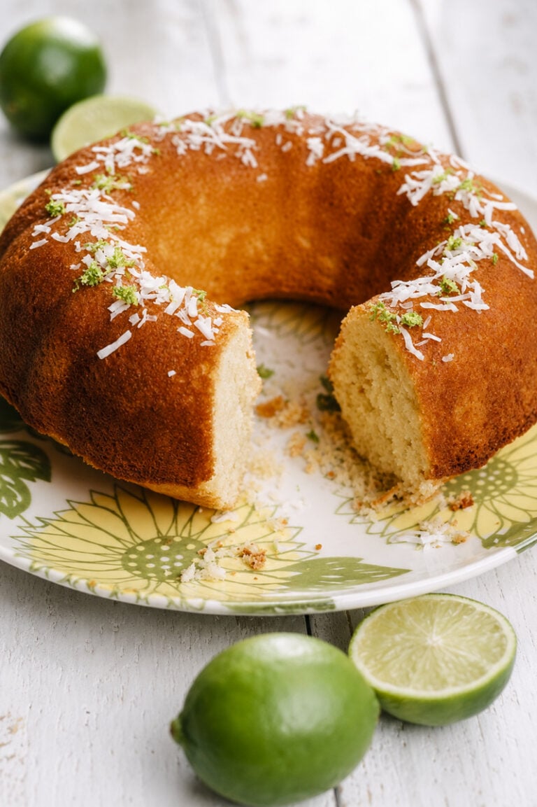 A Coconut Lime Bundt Cake topped with shredded coconut and lime zest sits on a floral plate. A slice is missing, and fresh limes are placed beside the cake on a white wooden surface.