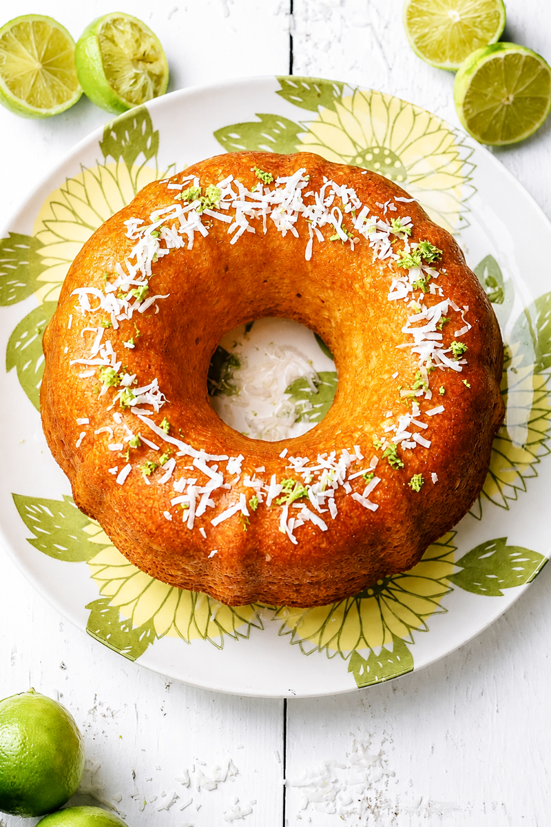 A Coconut Lime Bundt Cake, golden and moist, is topped with shredded coconut and lime zest as it sits on a floral plate, surrounded by halved and whole limes on a white wooden surface.