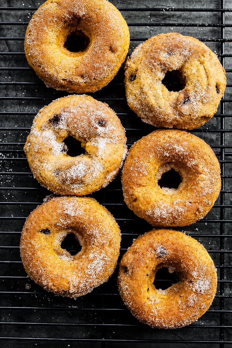 Six Vegan Pumpkin Chocolate Chip Donuts are cooling on a black wire rack, sugar-coated and arranged in two columns on a dark surface.
