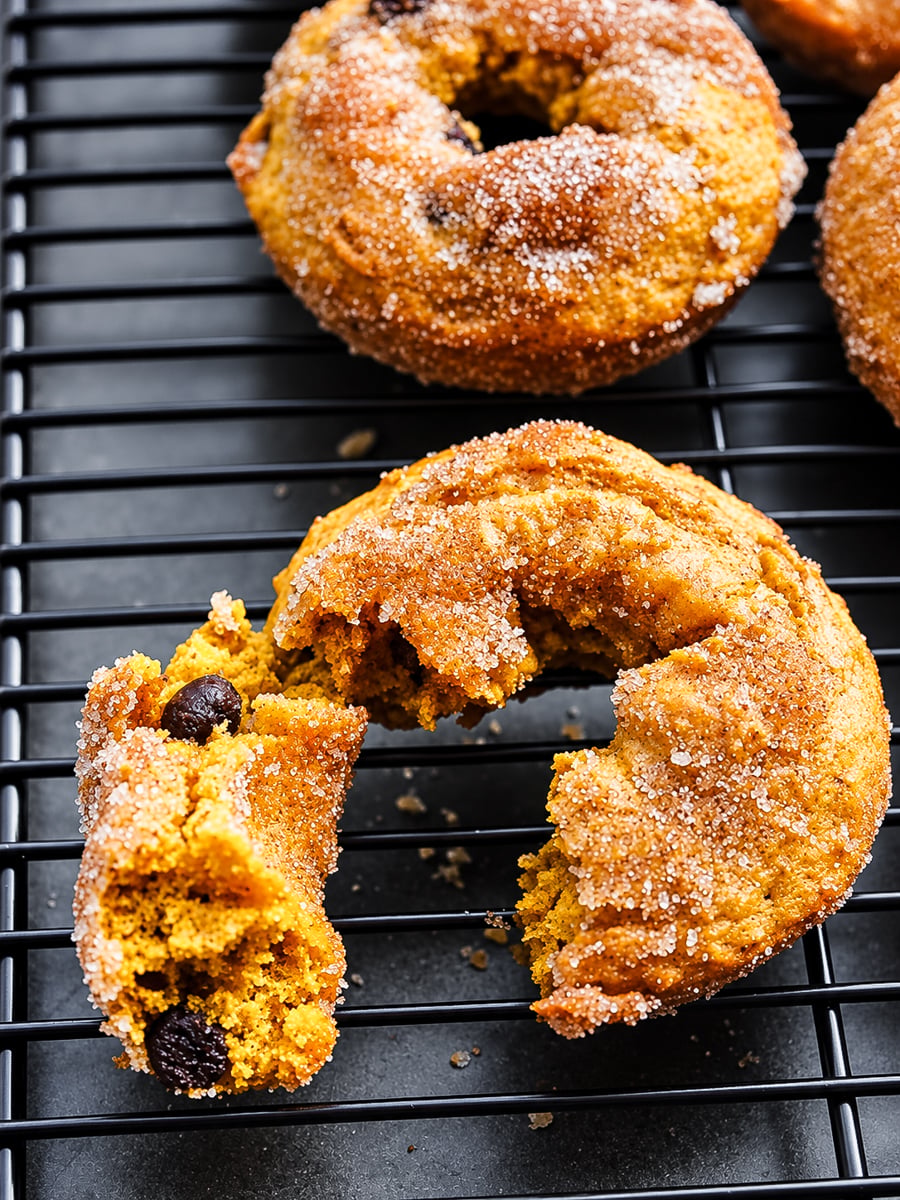 A Vegan Pumpkin Chocolate Chip Donut, broken in half, sits on a black cooling rack beside whole donuts.