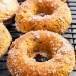 Three Vegan Pumpkin Chocolate Chip Donuts rest on a black cooling rack, boasting a golden-brown color and a light dusting of granulated sugar on their surfaces.