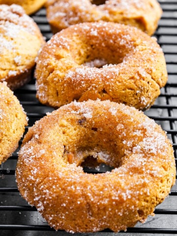 Three Vegan Pumpkin Chocolate Chip Donuts rest on a black cooling rack, boasting a golden-brown color and a light dusting of granulated sugar on their surfaces.