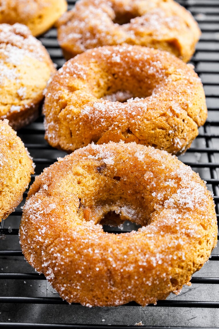 Three Vegan Pumpkin Chocolate Chip Donuts rest on a black cooling rack, boasting a golden-brown color and a light dusting of granulated sugar on their surfaces.