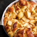 A round baking dish lined with parchment paper holds golden-brown Apple Harvest Bread pudding, with visible chunks of apples on top.