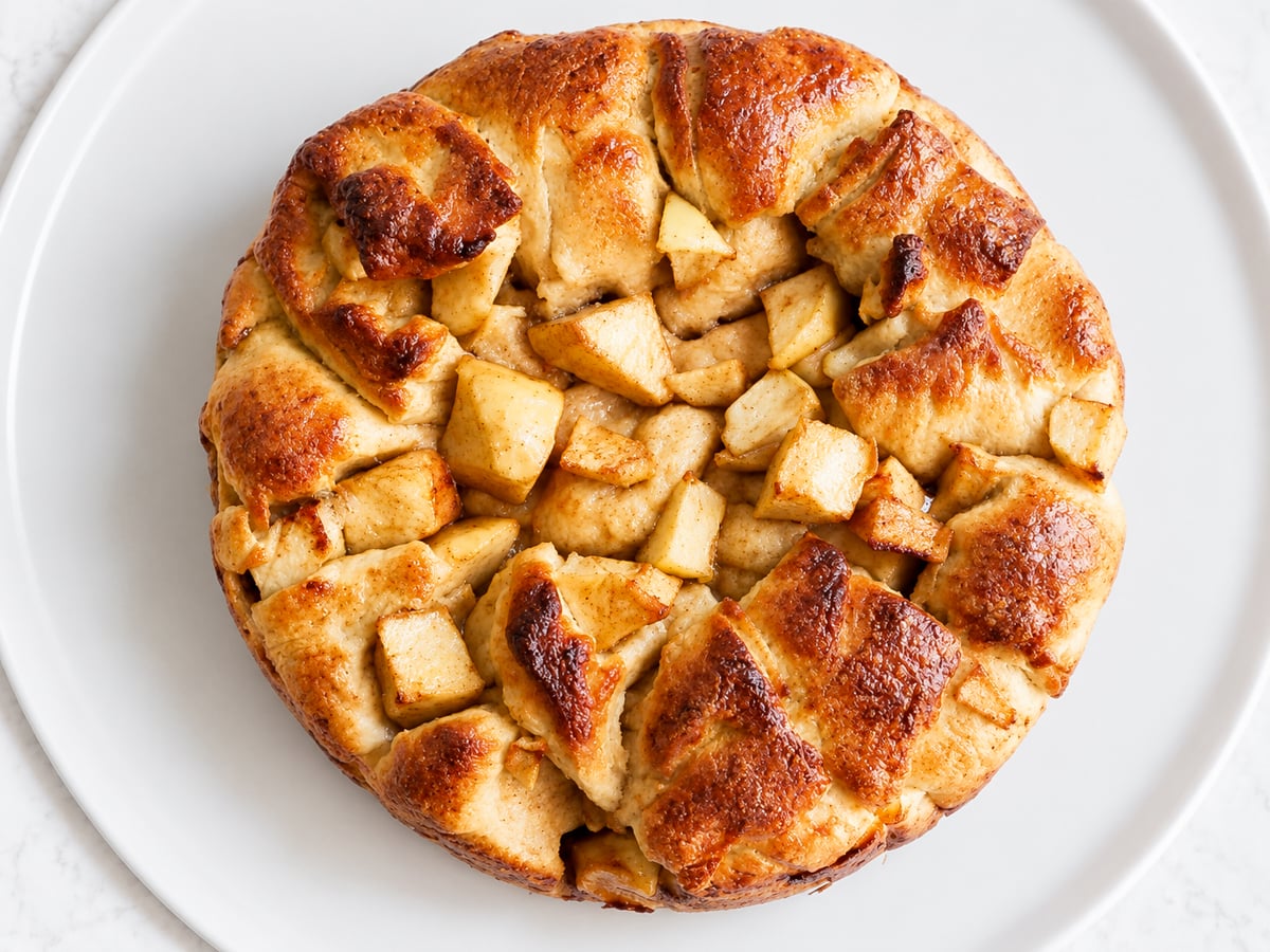 A round Apple Harvest Bread cake with golden-brown, rustic crust and visible baked apple chunks, served on a white plate.