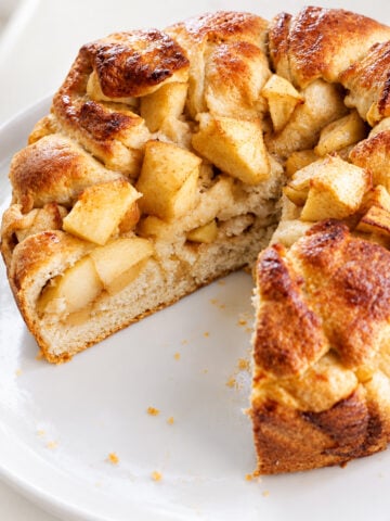 A round apple cake with a golden brown crust and chunks of apple, reminiscent of a classic Apple Harvest Bread, missing a slice and sitting on a white plate.