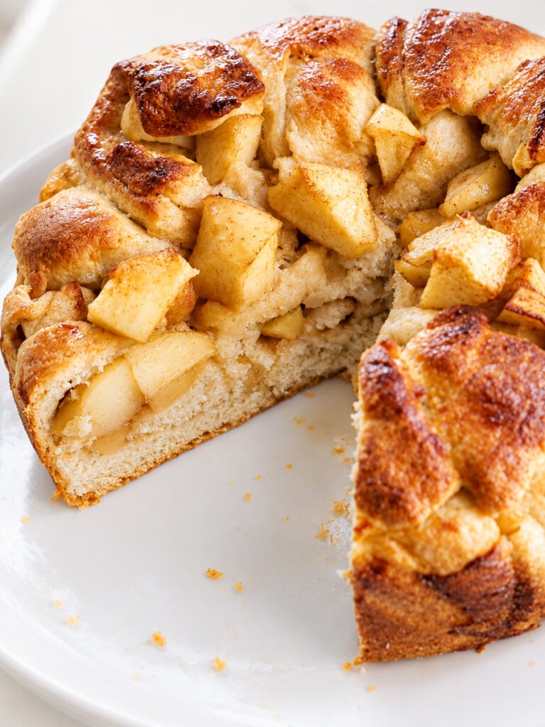 A round apple cake with a golden brown crust and chunks of apple, reminiscent of a classic Apple Harvest Bread, missing a slice and sitting on a white plate.