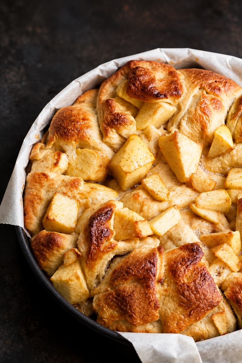 A round baking dish lined with parchment paper holds golden-brown Apple Harvest Bread pudding, with visible chunks of apples on top.