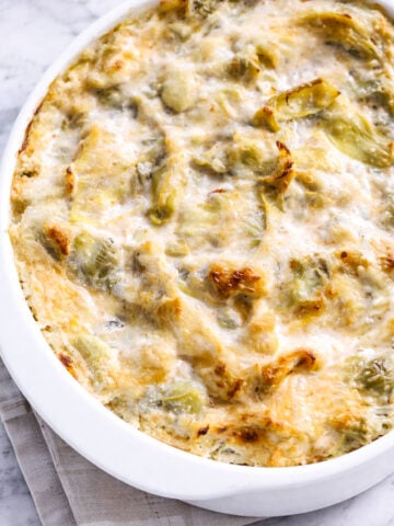 A hot and cheesy artichoke dip casserole with a golden, bubbly top and visible pieces of artichoke in a white oval dish, set on a folded beige cloth on a marble surface.