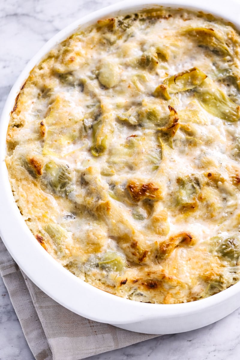 A hot and cheesy artichoke dip casserole with a golden, bubbly top and visible pieces of artichoke in a white oval dish, set on a folded beige cloth on a marble surface.