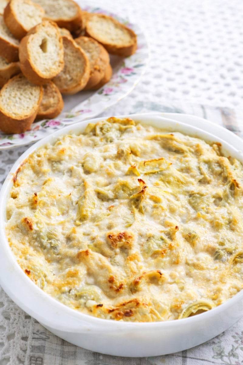 A round white dish filled with hot and cheesy artichoke dip, topped with melted cheese, sits on a lace tablecloth. In the background, a plate holds sliced toasted baguette pieces for dipping.