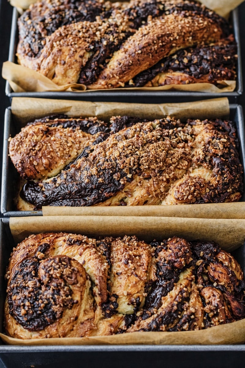 Three loaves of Chocolate Babka in parchment-lined pans, each with a swirled chocolate filling and crumbly streusel topping, are displayed close together.