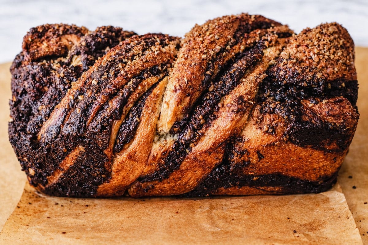 A close-up of a chocolate babka loaf with dark chocolate swirls and a crumbly topping, resting on a sheet of brown parchment paper.