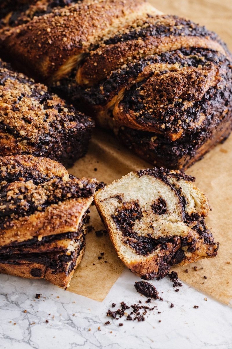 A sliced loaf of Chocolate Babka sits on parchment paper, revealing its swirled layers of chocolate and bread, topped with a crumbly streusel. One slice is separated from the loaf on a marble surface.