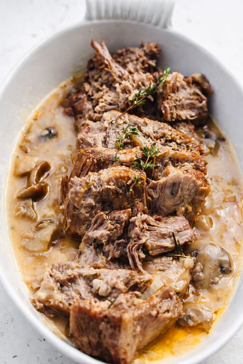 Sliced instant pot pork roast served in a white dish, topped with sprigs of fresh thyme and surrounded by a savory gravy with mushrooms and onions.