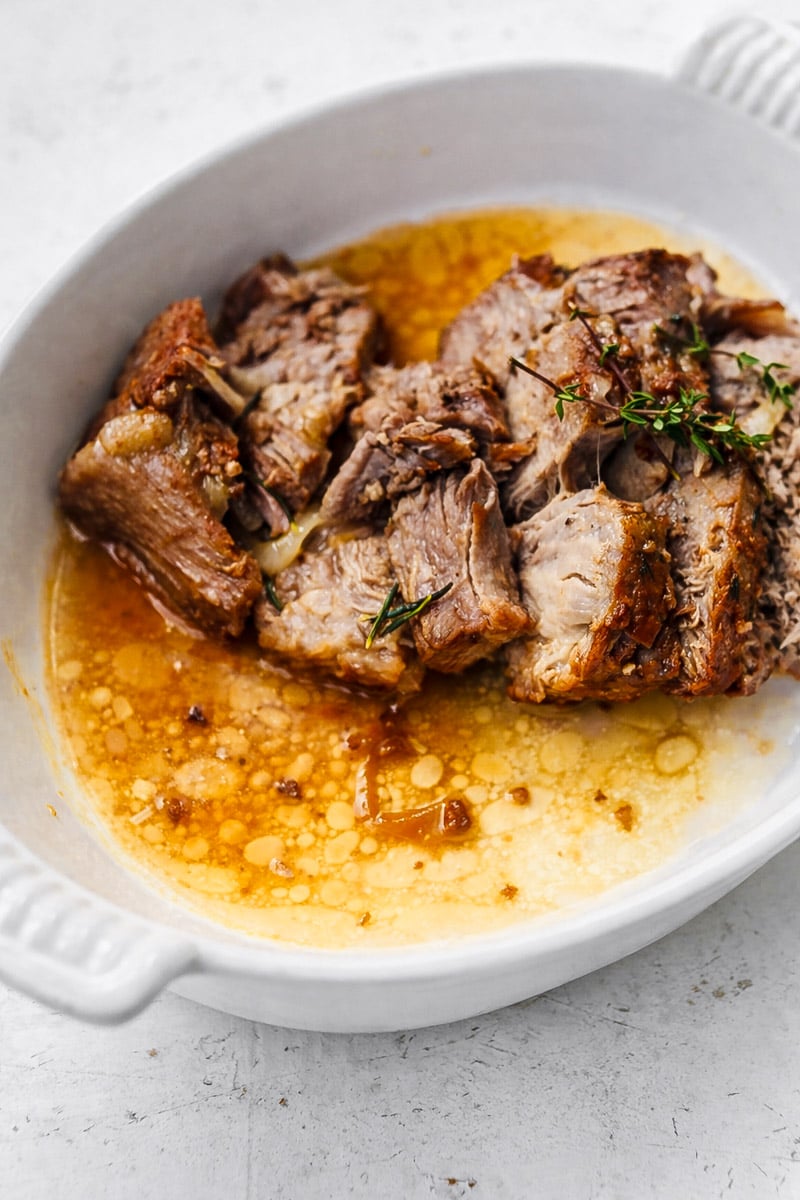 Tender pieces of Instant Pot pork roast sit in a white oval dish, surrounded by rich, golden pan juices and garnished with sprigs of fresh thyme.