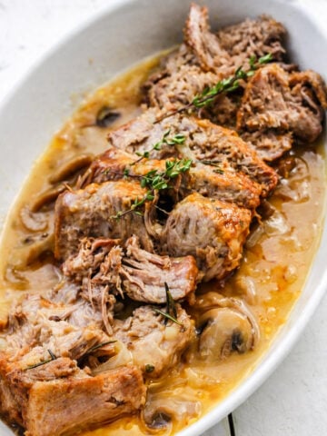 Slices of tender, braised beef pot roast served in a white oval dish with savory gravy-similar to an instant pot pork roast-garnished with sprigs of fresh thyme and visible pieces of mushrooms and onions.