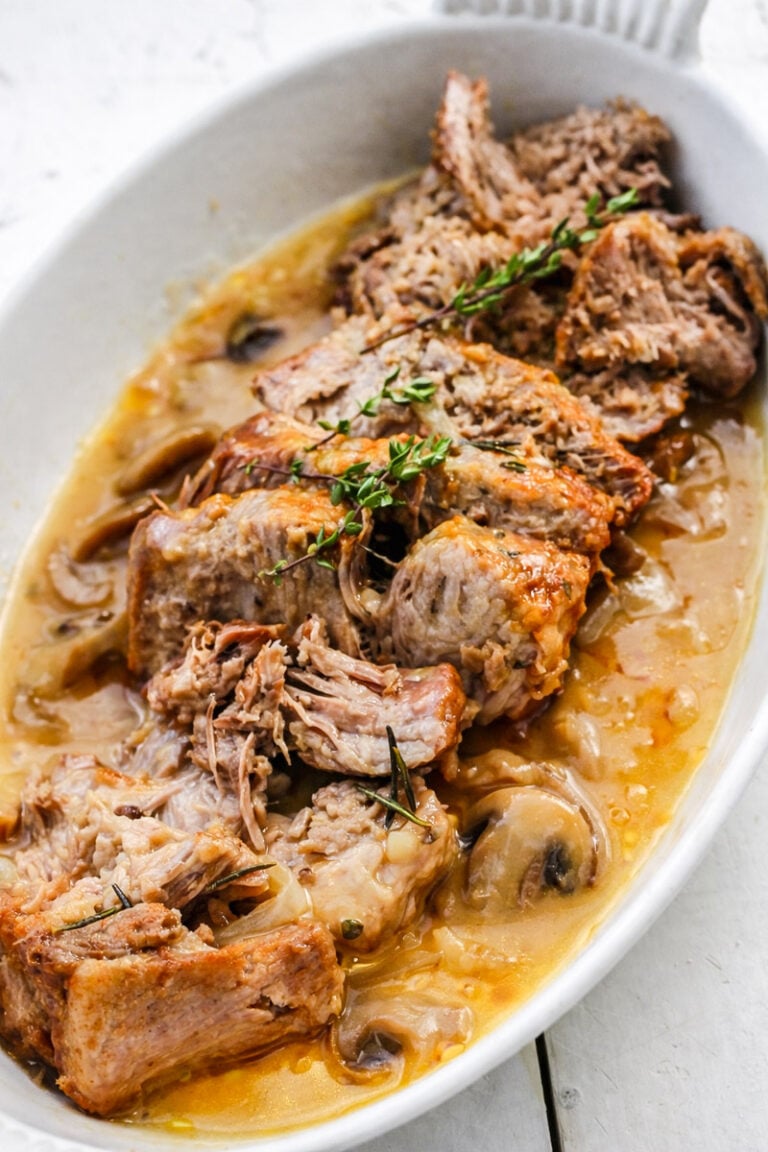 Slices of tender, braised beef pot roast served in a white oval dish with savory gravy—similar to an instant pot pork roast—garnished with sprigs of fresh thyme and visible pieces of mushrooms and onions.
