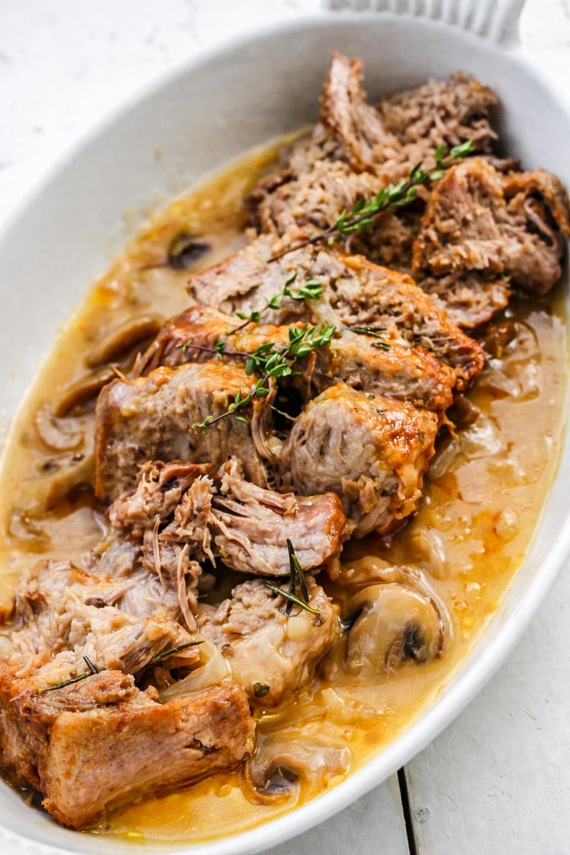 Slices of tender, braised beef pot roast served in a white oval dish with savory gravy-similar to an instant pot pork roast-garnished with sprigs of fresh thyme and visible pieces of mushrooms and onions.