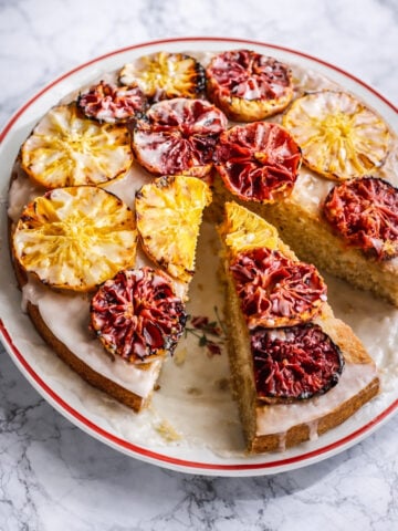 An Orange-Glazed Polenta Cake topped with vibrant, glazed slices of blood oranges and pineapples. One large slice is cut and slightly pulled out, showing the moist interior. The cake sits on a white plate with a red rim on a marble surface.