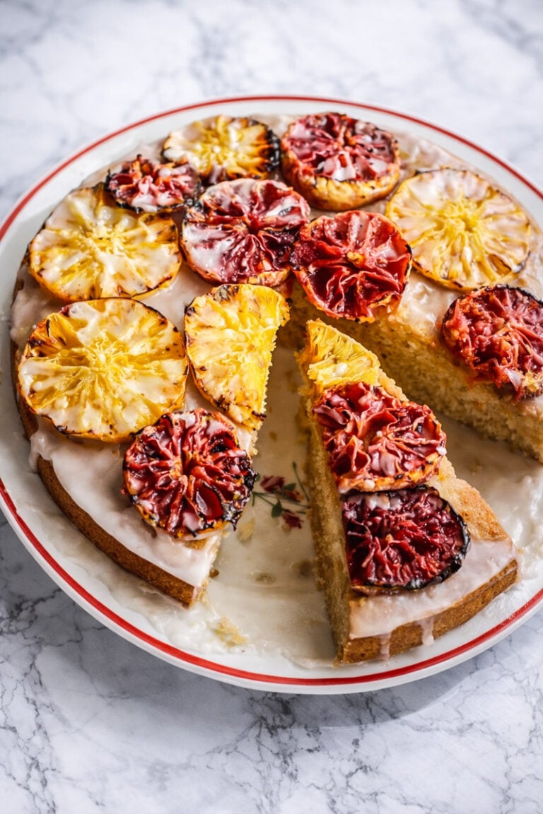 An Orange-Glazed Polenta Cake topped with vibrant, glazed slices of blood oranges and pineapples. One large slice is cut and slightly pulled out, showing the moist interior. The cake sits on a white plate with a red rim on a marble surface.