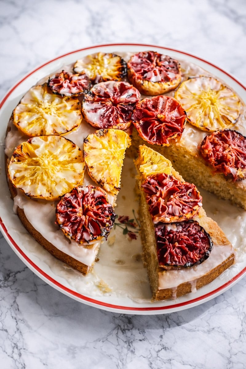 An Orange-Glazed Polenta Cake topped with vibrant, glazed slices of blood oranges and pineapples. One large slice is cut and slightly pulled out, showing the moist interior. The cake sits on a white plate with a red rim on a marble surface.