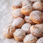 A pile of small round pastries, known as castagnole, dusted with powdered sugar sits on crumpled parchment paper, creating a cozy and inviting dessert scene.