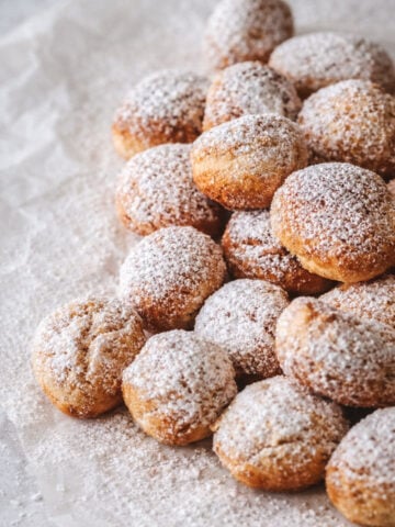 A pile of small round pastries, known as castagnole, dusted with powdered sugar sits on crumpled parchment paper, creating a cozy and inviting dessert scene.