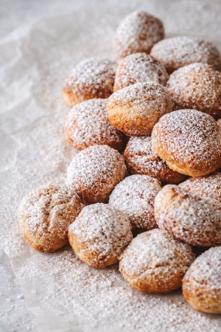 A pile of small round pastries, known as castagnole, dusted with powdered sugar sits on crumpled parchment paper, creating a cozy and inviting dessert scene.