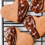 Four heart-shaped Espresso Shortbreads on a cooling rack-three are partially dipped in chocolate and sprinkled with coarse sea salt, while one cookie is plain without chocolate or salt.