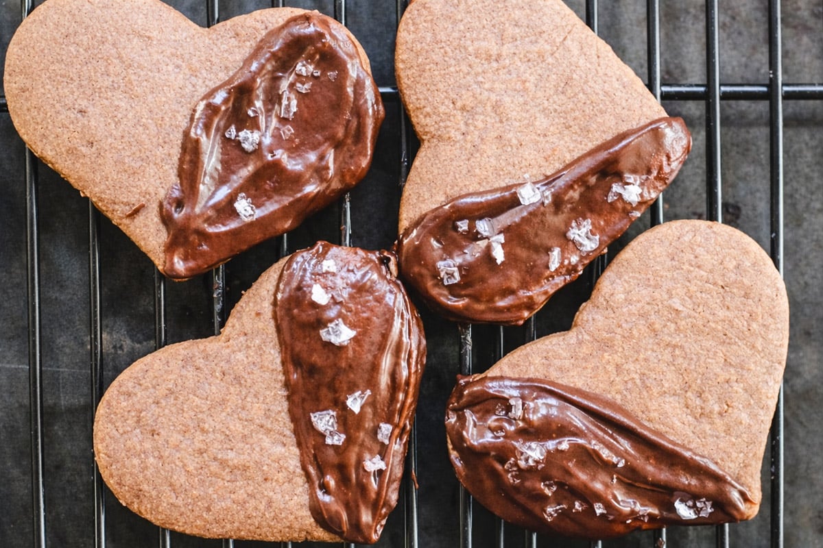 Four heart-shaped Espresso Shortbreads on a cooling rack, two partially coated with chocolate and sprinkled with sea salt, and two plain without chocolate.